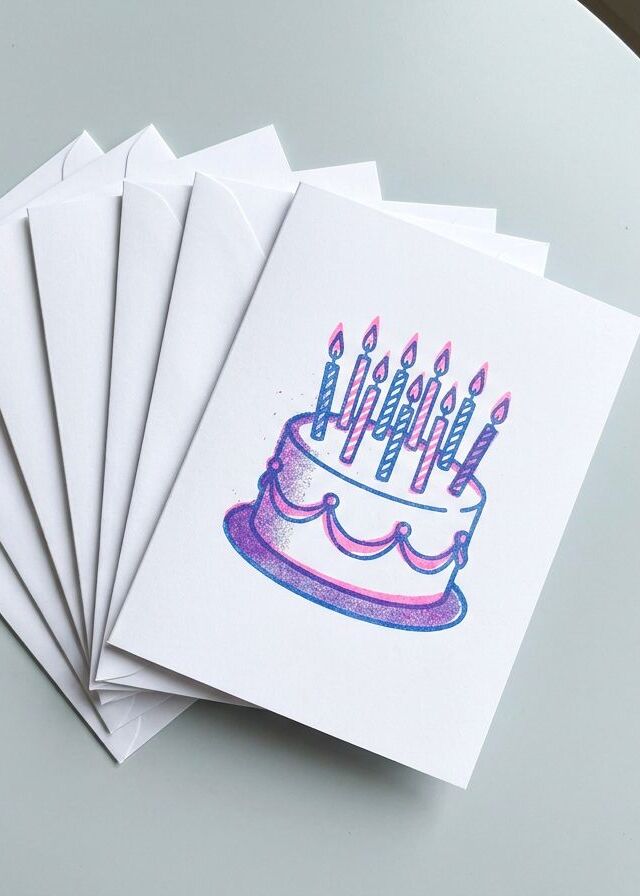A fan of printed birthday cards with a pink and blue cake, laid over white envelopes